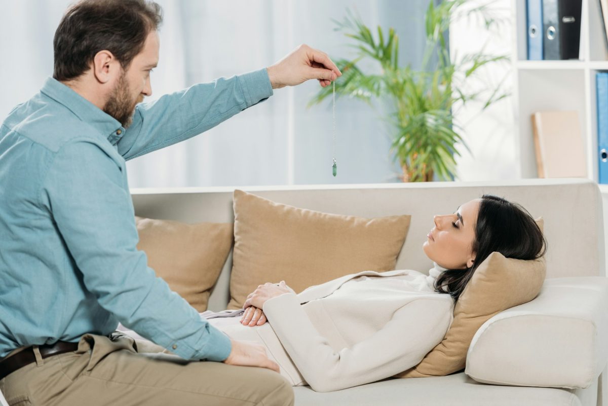 side view of bearded hypnotist holding pendulum and hypnotising young woman with closed eyes lying