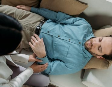 overhead view of hypnosis holding wrist of bearded man with closed eyes lying on couch